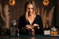 Self assured focused young female barkeeper with long blond hair in stylish outfit decorated cocktail with lemon slices while standing at counted in stylish bar - foto de stock