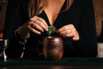 Обрезанная неузнаваемая самоуверенная молодая барменша с длинными светлыми волосами в стильном наряде, украшенном коктейлем с ломтиками лимона, стоя в стильном баре — стоковое фото