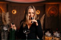 Задоволення молодої жінки барменом з довгим світлим волоссям, що пахне алкоголем, готуючи коктейль за стійкою — стокове фото