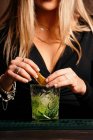 Cropped unrecognizable self assured focused young female barkeeper with long blond hair in stylish outfit decorated cocktail with lemon slices while standing at counted in stylish bar - foto de stock