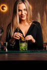 Self assured young female barkeeper with long blond hair in stylish outfit decorated cocktail with lemon slices while standing at counted in stylish bar — Foto stock