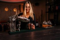 Schöne blonde junge Barkeeperin in stilvollem Outfit gießt Alkohol-Cocktail aus Shaker in eleganten Metallbecher im Restaurant — Stockfoto