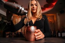 Schöne blonde junge Barkeeperin in stilvollem Outfit gießt Alkohol-Cocktail aus Shaker in eleganten Metallbecher im Restaurant — Stockfoto