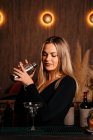 Красивая молодая барменша с длинными светлыми волосами в стильной одежде улыбается, смешивая коктейль в шейкере в современном баре — стоковое фото