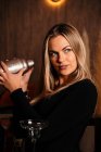 Belle jeune femme barman avec de longs cheveux blonds dans des vêtements élégants souriant tout en mélangeant cocktail dans shaker dans le bar moderne — Photo de stock