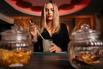 Blonde attraktive Barkeeperin rührt Eisalkohol-Cocktail mit langem Strohhalm auf Holztheke — Stockfoto