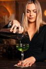 Schöne blonde junge Barkeeperin in stylischem Outfit gießt im Restaurant Alkohol-Cocktail aus Shaker in elegantes Margarita-Glas — Stockfoto