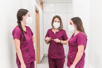 Group of young content nurses in medical uniform and masks having conversation in modern hospital corridor — Stock Photo