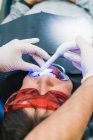 Dentiste anonyme de culture à angle élevé dans des gants à l'aide d'un outil de polymérisation aux ultraviolets dentaires pendant l'intervention avec le patient en clinique — Photo de stock
