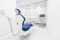 Интерьер современной аккуратной стоматологической клиники с синим стулом и белой мебелью, оборудованной современной стоматологической машиной и инструментами — стоковое фото