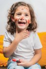Fröhliches kleines Mädchen in Freizeitkleidung mit roten Lippenstiftzeichnungen auf glücklichem Gesicht, das den Hals berührt und wegschaut, während es auf dem Boden sitzt — Stockfoto