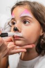 Crop artista creativo applicando vernici colorate corpo arte sul viso carino bambina in studio di luce — Foto stock