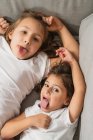 Сверху веселые маленькие сестры гримасуют и показывают языки, лежа на удобном диване и глядя в камеру — стоковое фото