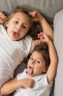 From above cheerful little sisters grimacing and showing tongues while lying on comfy couch and looking at camera — Stock Photo