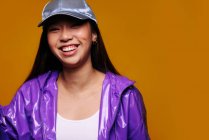 Retrato da jovem asiática feliz. Ela usa uma jaqueta roxa e um boné cinza e está olhando para a câmera sorrindo contra um fundo amarelo — Fotografia de Stock