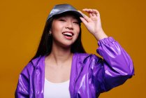 Портрет щасливої азіатської дівчини. Вона носить пурпуровий піджак і сіру шапку і дивиться на камеру, посміхаючись на жовтому тлі — стокове фото