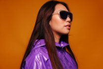 Retrato de una joven asiática con expresión seria. Lleva una chaqueta morada y unas gafas de sol negras y está mirando hacia otro lado con un fondo amarillo. - foto de stock