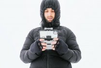 Focado barbudo masculino vestindo casaco preto quente com capuz em pé em terreno nevado com controle remoto drone e olhando para longe — Fotografia de Stock
