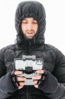 Homme barbu concentré portant une veste noire chaude avec capuche debout sur un terrain enneigé avec télécommande par drone et détournant les yeux — Photo de stock