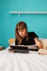 Концентрированная молодая женщина с татуированной рукой просматривает современный планшет и отдыхает на удобной кровати в светлой спальне — стоковое фото