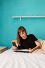 Mujer joven concentrada con brazo tatuado que navega por la tableta moderna y descansa en una cama cómoda en un dormitorio luminoso - foto de stock