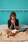 Serene female in casual black shirt with tattooed arms enjoying cup of tea and sitting on cozy bed with eyes near modern tablet and adorable sleeping dog — Stock Photo
