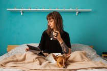 Jovem mulher positiva em camisa preta desfrutando de chá quente e assistindo vídeo em tablet enquanto descansa na cama confortável com adorável cão amigável — Fotografia de Stock