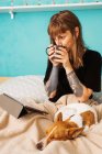 Позитивна молода жінка в чорній сорочці насолоджується гарячим чаєм і дивиться відео на планшет, відпочиваючи на зручному ліжку з чарівною доброзичливою собакою — стокове фото