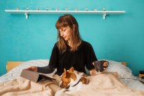 Positive junge Frau im schwarzen Hemd genießt heißen Tee und schaut sich Videos auf dem Tablet an, während sie sich auf einem bequemen Bett mit einem entzückenden freundlichen Hund ausruht — Stockfoto