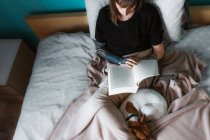 Зверху врожаю зосереджена жінка в чорній сорочці і татуйована рука відпочиває на затишному ліжку з милою сплячою собакою і читає цікаву книгу — стокове фото