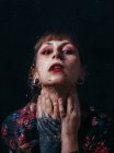 Mulher sem emoção com braço tatuagem tocando pescoço em pé atrás de vidro translúcido com gotas de água olhando para a câmera — Fotografia de Stock
