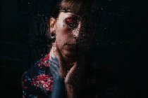 Crop chateado fêmea com olhos fechados e braço pintado tocando pescoço atrás de vidro translúcido com gotas de água — Fotografia de Stock