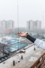 Cultiver une personne anonyme attrapant des flocons de neige tombants tout en se tenant sur le balcon dans la ville moderne par une froide journée d'hiver — Photo de stock