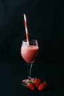 Strömung von leckeren Getränken, die in transparentes Glas mit gestreiftem Stroh neben saftigen Erdbeeren auf schwarzem Hintergrund gegossen werden — Stockfoto