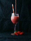 Flow of yummy beverage pouring into transparent glass with striped straw near juicy strawberries on black background — Stock Photo