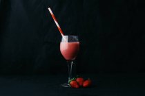 Flujo de deliciosa bebida vertiendo en vidrio transparente con paja rayada cerca de fresas jugosas sobre fondo negro - foto de stock
