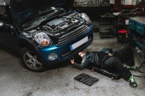 Junger Autoservice-Mann kontrolliert Fahrzeug mit offener Motorhaube, während er in Werkstatt auf Matte liegt — Stockfoto