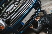 Junger Autoservice-Mann kontrolliert Fahrzeug mit offener Motorhaube, während er in Werkstatt auf Matte liegt — Stockfoto