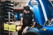 Молодой сфокусированный бородатый ремонтник ремонтирует современный автомобиль против яркого света и смотрит в сторону в мастерской — стоковое фото
