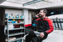 Mestre masculino com netbook falando no celular perto de transporte na garagem — Fotografia de Stock