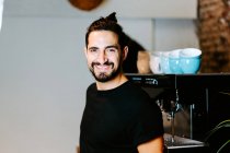 Smiling male barista using portafilter and preparing coffee in modern coffeemaker while standing at counter in cafe and looking at camera — Stock Photo