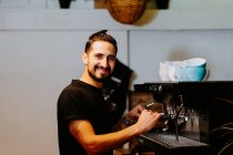 Vista lateral de sorrir barista masculino usando portafilter e preparar café na cafeteira moderna, enquanto está em pé no balcão no café e olhando para a câmera — Fotografia de Stock