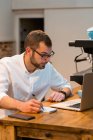 Konzentrierter männlicher Barista sitzt an Holztheke mit Laptop und macht sich Notizen in Notizbuch, während er im Café arbeitet — Stockfoto