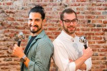 Side view of cheerful male baristas with portafilters standing back to back and looking at camera on background of brick wall in loft styled cafe — Stock Photo