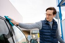 Focused young male in casual clothes and eyeglasses wiping vehicle with rag while standing in car wash station against cloudy sky — Stock Photo