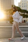 Side view of female wearing protective mask walking along city street and browsing smartphone during coronavirus pandemic — Stock Photo