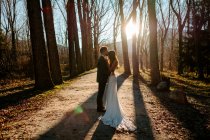 Vista lateral do noivo jovem irreconhecível no terno elegante beijando a testa da noiva graciosa no vestido branco enquanto estava na floresta de outono no dia do casamento — Fotografia de Stock