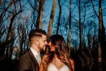Side view of young ethnic bride with long hair in white dress smiling and hugging groom with closed eyes while standing together near coniferous tree in forest — Stock Photo