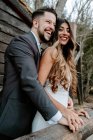 Боковой вид веселый молодой бородатый жених в костюме смеется и обнимает элегантную этническую невесту в белом платье, стоя вместе на деревянном мосту возле старого дома в осеннем лесу — стоковое фото