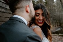 Seitenansicht des jungen Bräutigams im eleganten Anzug küsst lächelnde ethnische Braut, während sie am Hochzeitstag in der Natur steht — Stockfoto